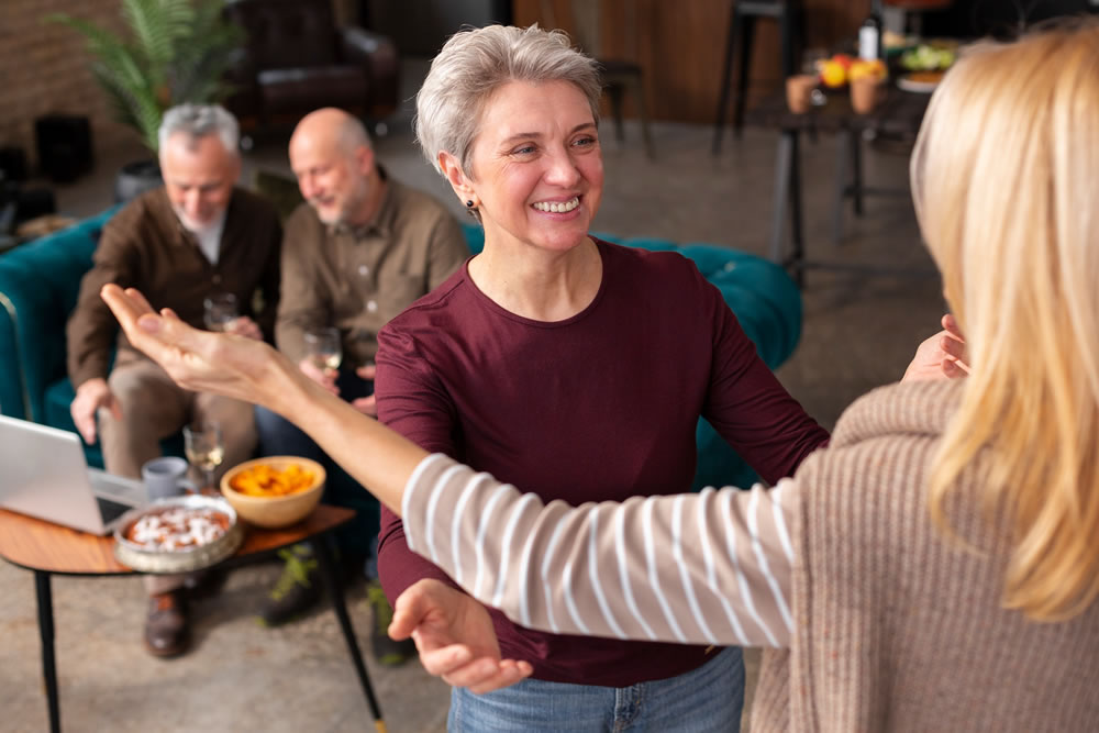 Mujer mayor sonriendo y saludando con los brazos abiertos a otra persona en una reunión social con amigos al fondo